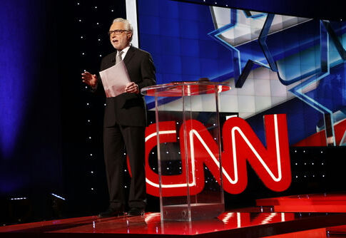 news anchor stands on stage beside a clear podium