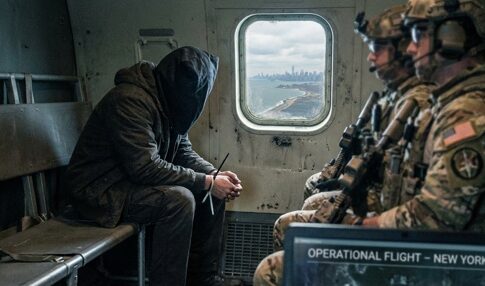 A hooded figure sits quietly in a helicopter with soldiers, overlooking a city skyline.